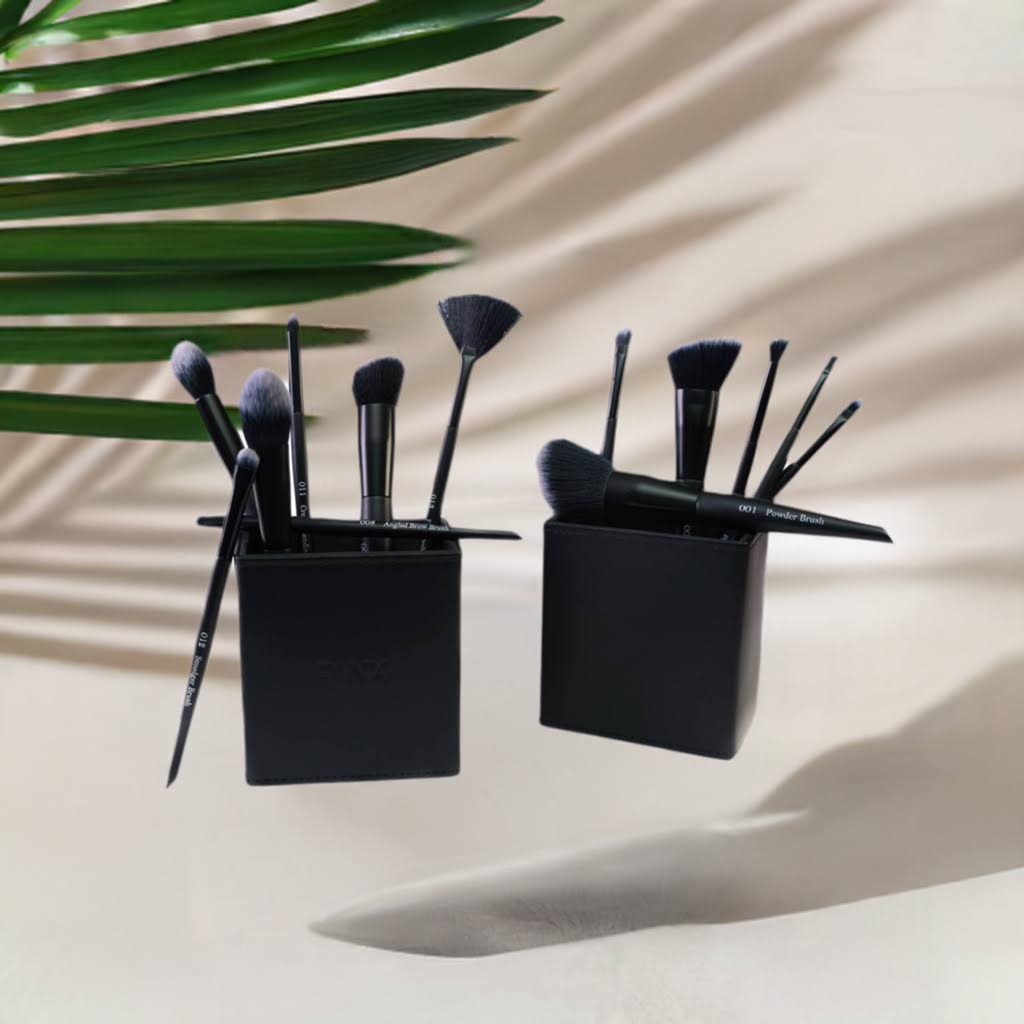 A set of professional makeup brushes with matte black handles and wooden handles, displayed in a shadowed setting, with a palm leaf in the background.